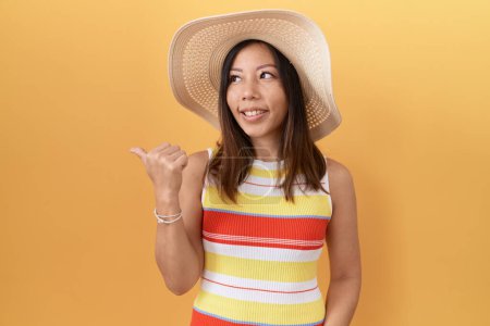 Téléchargez les photos : Femme chinoise du Moyen Âge portant un chapeau d'été sur fond jaune souriant avec un visage heureux regardant et pointant vers le côté avec le pouce vers le haut. - en image libre de droit