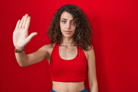 Photo for Hispanic woman with curly hair standing over red background doing stop sing with palm of the hand. warning expression with negative and serious gesture on the face. - Royalty Free Image
