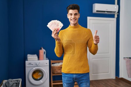 Foto de Joven hombre hispano en la lavandería sosteniendo shekels sonriendo feliz y positivo, pulgar hacia arriba haciendo excelente y signo de aprobación - Imagen libre de derechos