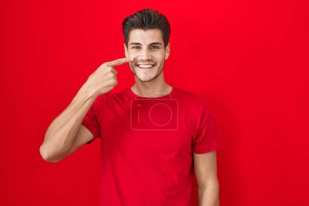 Téléchargez les photos : Jeune homme hispanique debout sur fond rouge pointant avec le doigt de la main sur le visage et le nez, souriant gai. concept de beauté - en image libre de droit