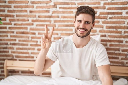 Téléchargez les photos : Young caucasian man doing victory gesture sitting on bed at bedroom - en image libre de droit