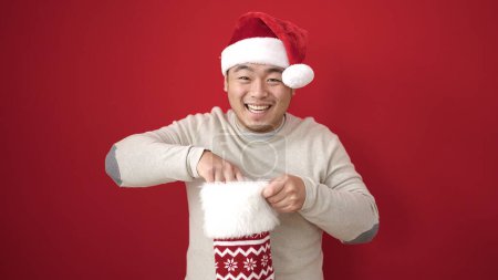 Téléchargez les photos : Jeune homme chinois souriant confiant tenant chaussette de Noël sur fond rouge isolé - en image libre de droit