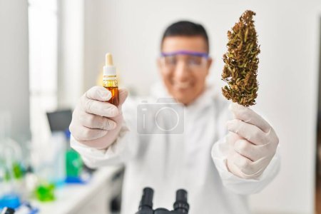 Téléchargez les photos : Jeune homme latin scientifique tenant de l'herbe et du liquide de cannabis en laboratoire - en image libre de droit