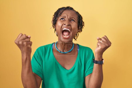 Photo for African woman with dreadlocks standing over yellow background angry and mad screaming frustrated and furious, shouting with anger. rage and aggressive concept. - Royalty Free Image