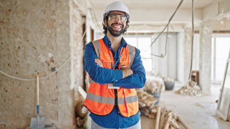 Téléchargez les photos : Jeune homme hispanique architecte souriant confiant debout les bras croisés geste sur le chantier de construction - en image libre de droit