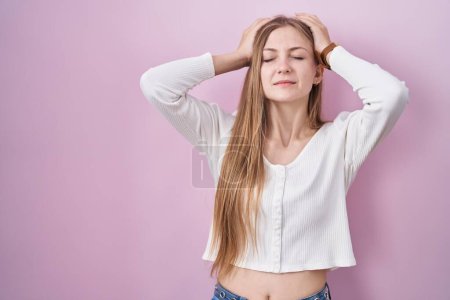 Foto de Young caucasian woman standing over pink background suffering from headache desperate and stressed because pain and migraine. hands on head. - Imagen libre de derechos