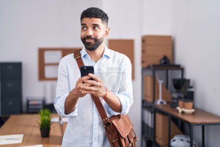 Téléchargez les photos : Homme hispanique avec la barbe à l'aide d'un smartphone au bureau souriant regardant vers le côté et regardant loin en pensant. - en image libre de droit