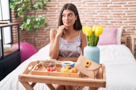 Photo for Brunette young woman eating breakfast sitting on the bed serious face thinking about question with hand on chin, thoughtful about confusing idea - Royalty Free Image