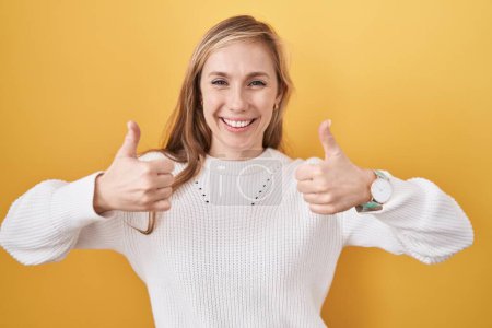Foto de Mujer joven caucásica con suéter blanco sobre fondo amarillo signo de éxito haciendo gesto positivo con la mano, pulgares hacia arriba sonriendo y feliz. expresión alegre y gesto ganador. - Imagen libre de derechos