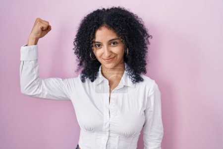 Téléchargez les photos : Femme hispanique avec les cheveux bouclés debout sur fond rose personne forte montrant muscle du bras, confiant et fier de la puissance - en image libre de droit