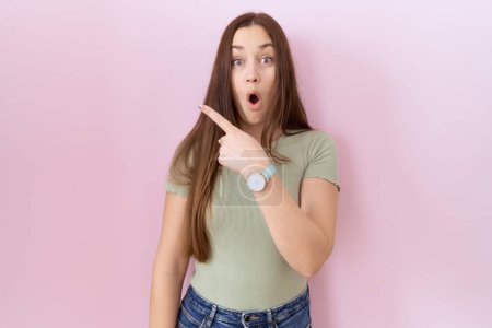 Photo for Beautiful brunette woman standing over pink background surprised pointing with finger to the side, open mouth amazed expression. - Royalty Free Image