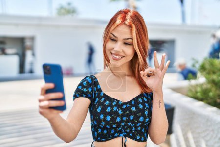 Photo for Young caucasian woman doing video call with smartphone doing ok sign with fingers, smiling friendly gesturing excellent symbol - Royalty Free Image