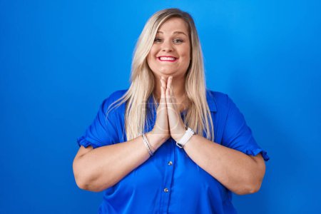 Photo for Caucasian plus size woman standing over blue background praying with hands together asking for forgiveness smiling confident. - Royalty Free Image
