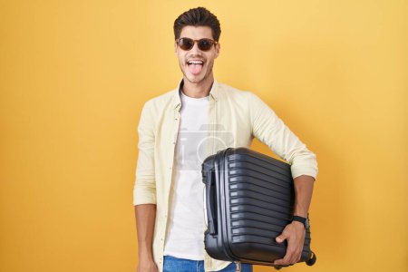 Photo for Young hispanic man holding suitcase going on summer vacation sticking tongue out happy with funny expression. emotion concept. - Royalty Free Image