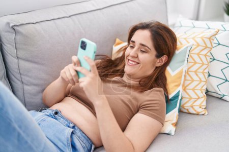 Téléchargez les photos : Jeune femme utilisant un smartphone couché sur le canapé à la maison - en image libre de droit