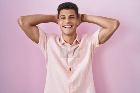 Foto de Joven hombre hispano de pie sobre fondo rosa relajante y estiramiento, brazos y manos detrás de la cabeza y el cuello sonriendo feliz - Imagen libre de derechos