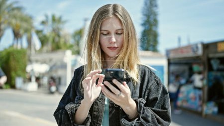 Téléchargez les photos : Jeune femme blonde utilisant un smartphone avec un visage sérieux à la rue - en image libre de droit