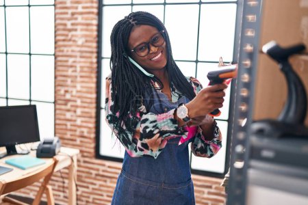 Foto de Mujer afroamericana comercio electrónico trabajador de negocios paquete de escaneo hablando en el teléfono inteligente en la oficina - Imagen libre de derechos