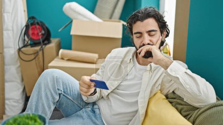 Photo for Young hispanic man holding credit card sitting on sofa thinking at new home - Royalty Free Image