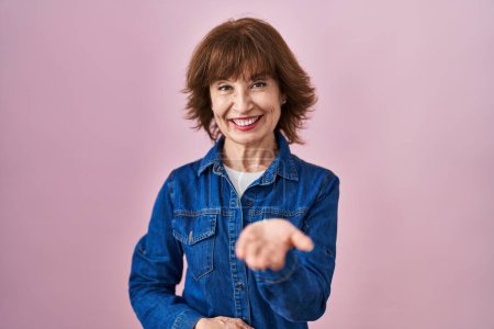 Foto de Mujer de mediana edad de pie sobre fondo rosa sonriente alegre ofreciendo la palma de la mano dando asistencia y aceptación. - Imagen libre de derechos