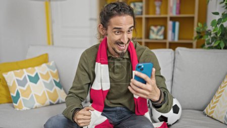 Foto de Hombre hispano joven viendo el partido de fútbol en el teléfono inteligente de apoyo en casa - Imagen libre de derechos