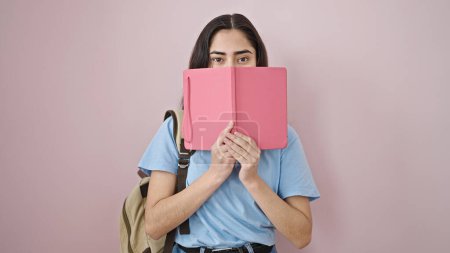 Foto de Joven hermosa mujer hispana estudiante cubriendo la boca con libro sobre fondo rosa aislado - Imagen libre de derechos