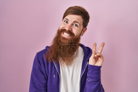 Téléchargez les photos : Homme caucasien avec une longue barbe debout sur fond rose souriant avec un sourire heureux clin d'oeil à la caméra faisant signe de victoire. numéro deux. - en image libre de droit
