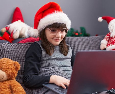 Téléchargez les photos : Adorable fille hispanique portant un chapeau de Noël en utilisant un ordinateur portable à la maison - en image libre de droit
