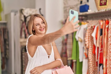 Téléchargez les photos : Jeune femme blonde client tenant des sacs à provisions faire selfie par smartphone au magasin de vêtements - en image libre de droit