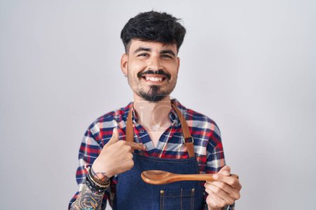Foto de Joven hombre hispano con barba llevando delantal degustación de comida sosteniendo cuchara de madera señalando con el dedo a uno mismo sonriendo feliz y orgulloso - Imagen libre de derechos