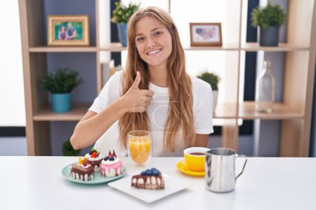 Photo for Young caucasian woman eating pastries t for breakfast doing happy thumbs up gesture with hand. approving expression looking at the camera showing success. - Royalty Free Image