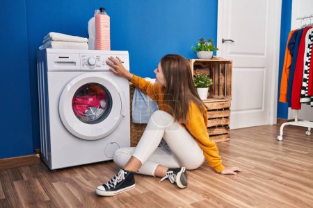 Photo for Young blonde woman sitting on floor turning washing machine on at laundry room - Royalty Free Image