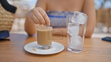 Photo for Young hispanic woman stirring coffee sitting on the table at sunny restaurant terrace - Royalty Free Image