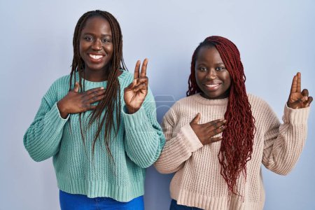 Téléchargez les photos : Deux femmes africaines debout sur fond bleu souriant jurant avec la main sur la poitrine et les doigts vers le haut, faisant un serment de fidélité - en image libre de droit
