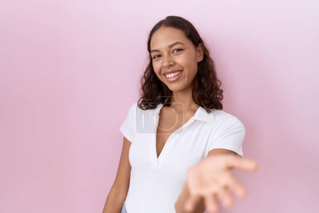 Foto de Mujer hispana joven vistiendo camiseta blanca casual sonriente alegre ofreciendo mano de palma dando asistencia y aceptación. - Imagen libre de derechos