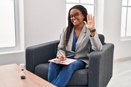 Téléchargez les photos : Jeune afro-américain avec des tresses travaillant au bureau de consultation renonçant à dire bonjour heureux et souriant, geste de bienvenue amical - en image libre de droit