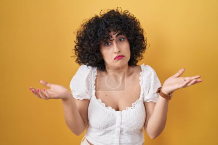 Photo for Young brunette woman with curly hair standing over yellow background clueless and confused expression with arms and hands raised. doubt concept. - Royalty Free Image