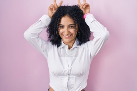 Téléchargez les photos : Femme hispanique avec les cheveux bouclés debout sur fond rose posant drôle et fou avec les doigts sur la tête comme des oreilles de lapin, souriant gai - en image libre de droit