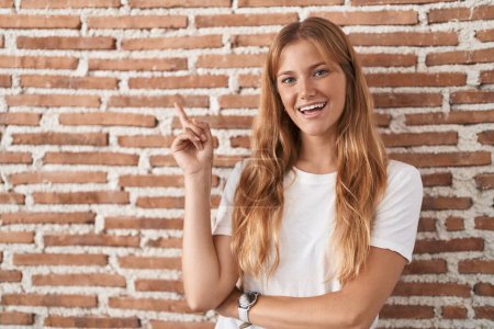 Foto de Mujer caucásica joven de pie sobre la pared de ladrillos con una gran sonrisa en la cara, señalando con el dedo de la mano al lado mirando a la cámara. - Imagen libre de derechos