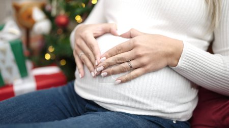 Photo for Young pregnant woman celebrating christmas touching belly doing heart gesture at home - Royalty Free Image