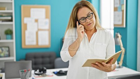 Foto de Trabajadora de negocios hispana de mediana edad hablando por teléfono leyendo cuaderno en la oficina - Imagen libre de derechos