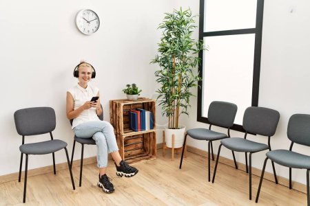 Foto de Mujer rubia joven usando teléfono inteligente y auriculares sentados en la silla en la sala de espera - Imagen libre de derechos