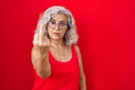 Foto de Middle age woman with grey hair standing over red background showing middle finger, impolite and rude fuck off expression - Imagen libre de derechos