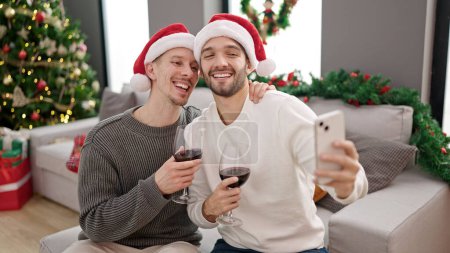 Foto de Dos hombres pareja celebrando la Navidad bebiendo vino teniendo videollamada en casa - Imagen libre de derechos