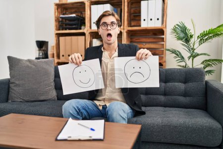 Photo for Young therapist man working on depression holding sad to happy emotion paper afraid and shocked with surprise and amazed expression, fear and excited face. - Royalty Free Image