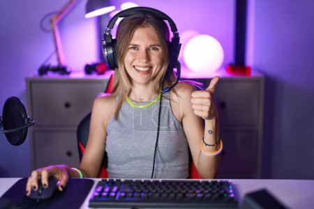 Téléchargez les photos : Blonde femme caucasienne jouer à des jeux vidéo avec casque souriant heureux et positif, pouce levé faire excellent et signe d'approbation - en image libre de droit