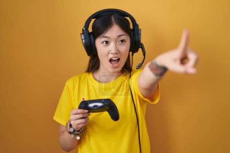 Foto de Chino joven mujer jugando videojuego celebración controlador apuntando con el dedo sorprendido por delante, boca abierta expresión asombrada, algo en el frente - Imagen libre de derechos