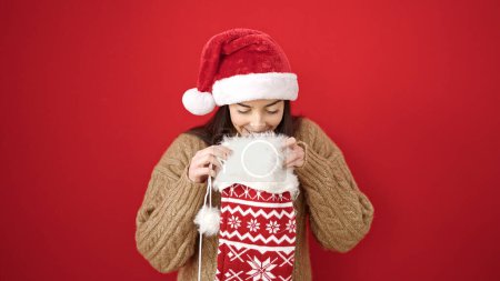 Téléchargez les photos : Jeune belle femme hispanique portant un chapeau de Noël regardant à l'intérieur de la chaussette sur fond rouge isolé - en image libre de droit