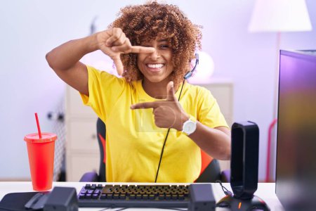 Foto de Mujer hispana joven con el pelo rizado jugando videojuegos con auriculares sonriendo haciendo marco con las manos y los dedos con la cara feliz. concepto de creatividad y fotografía. - Imagen libre de derechos