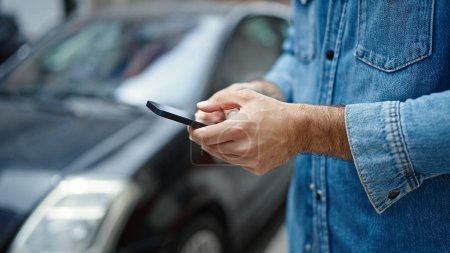 Téléchargez les photos : Jeune homme hispanique utilisant smartphone debout en voiture à la rue - en image libre de droit